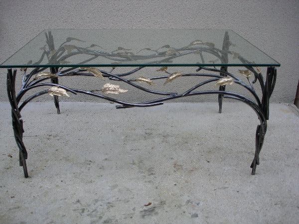 table basse avec piètement en acier ; un bra,chage en acier crée un décor sous le plateau de verre ; il est agrémenté de feuilles de laiton. vue de profil