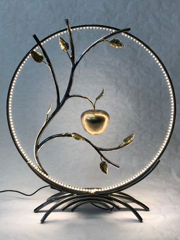 lampe à poser composée d'un cercle d'acier dans lequel un branchage avec feuilles de laiton abrite une pomme martenée en volume ; la pomme est en laiton ; un ruban led fait le pourtour du cercle d'acier ; lampe allumée sur fond blanc