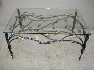 table basse avec piètement en acier ; un bra,chage en acier crée un décor sous le plateau de verre ; il est agrémenté de feuilles de laiton. vue de dessus