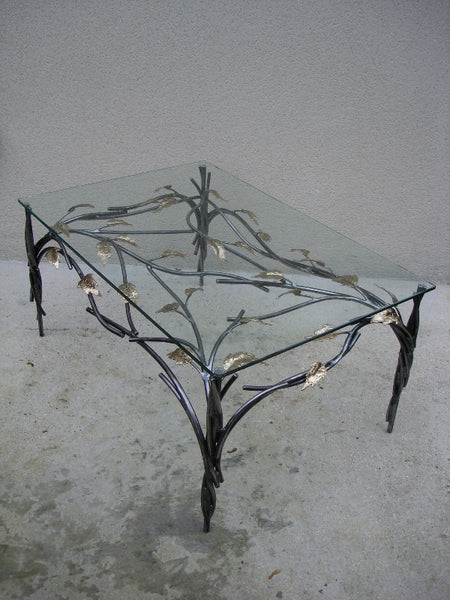 table basse avec piètement en acier ; un bra,chage en acier crée un décor sous le plateau de verre ; il est agrémenté de feuilles de laiton. vue de dessus et de côté