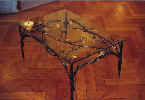 table basse avec piètement en acier ; un bra,chage en acier crée un décor sous le plateau de verre ; il est agrémenté de feuilles de laiton. vue de biais sur parquet